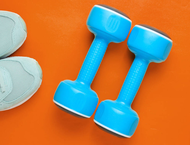 Dwa niebieskie plastikowe hantle, buty sportowe na pomarańczowym tle Sport, koncepcja fitness. Widok góry. Minimalizm - Zdjęcie, obraz