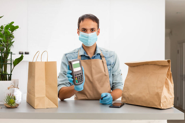 delivery man in a protective mask and gloves holds pos terminal in hand. Service quarantine pandemic coronavirus virus concept - Φωτογραφία, εικόνα