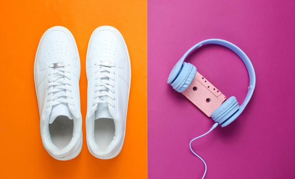 Retro-Stil. 80er Jahre. Popkultur. Minimalismus. Kopfhörer mit Audiokassette, weiße Turnschuhe vor lila-orangefarbenem Hintergrund. Ansicht von oben - Foto, Bild