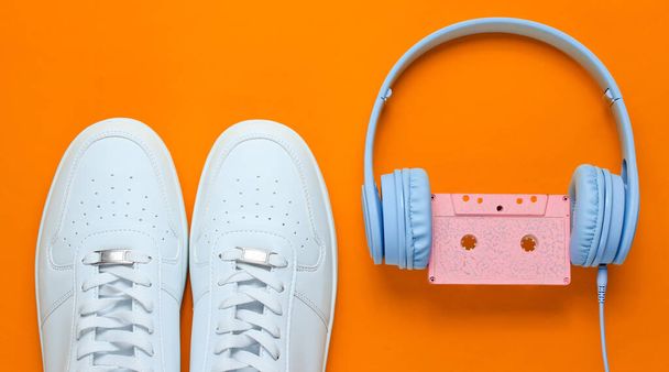 Retro tarzı. 80 'ler. Popüler kültür. Minimalizm. Ses kaseti olan kulaklıklar, turuncu arka planda beyaz spor ayakkabılar. Üst görünüm - Fotoğraf, Görsel