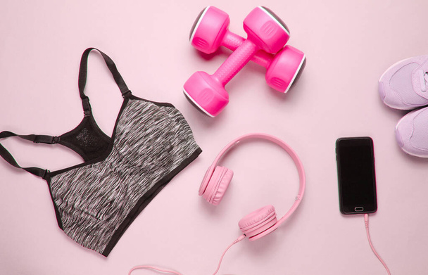 Фитнес-концепция. Плоская укладка смартфона с наушниками, пластиковыми гантели, спортивный бюстгальтер, кроссовки на розовом фоне. Вид сверху - Фото, изображение