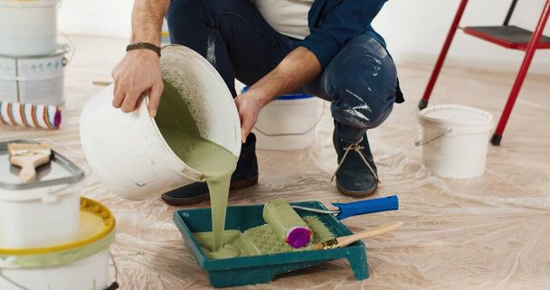 Close up of Caucasian young handsome man pouring green olive paint preparing for painting walls in house renovating and redesigning room. Handyman working on apartment redecoration repair and makeover - Photo, Image