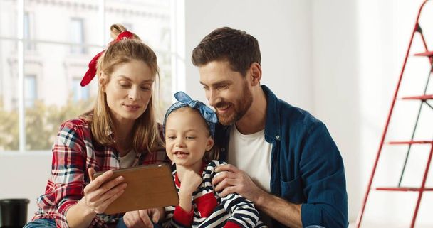 Joyful Caucasian parents sitting with little cute preschool girl chatting, tapping and browsing online on tablet device, choosing new room decor on internet together. Home repair concept - Fotoğraf, Görsel