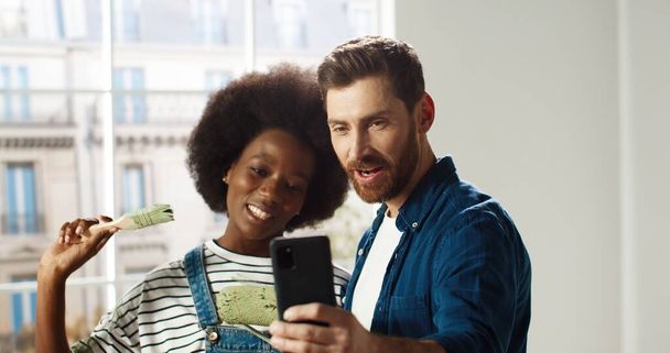 Portrait of happy mixed-races young married family couple man and woman smiling in new apartment taking selfie photos on smartphone posing with brushes in room. Home repair concept - Foto, imagen