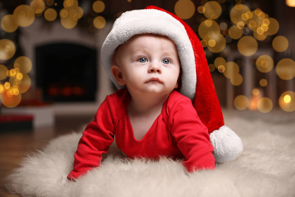 Cute little baby in red pajamas and Santa hat on floor against blurred festive lights. Christmas suit - Photo, Image