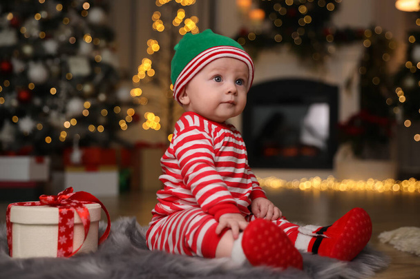Cute little baby wearing Santa's elf clothes with Christmas gift on floor at home - Foto, Imagem