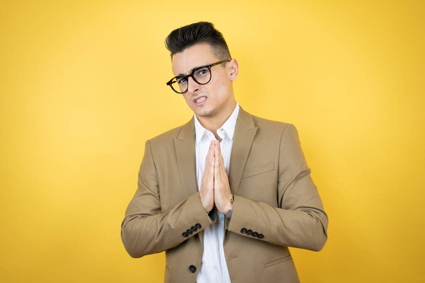 Молодой бизнесмен на изолированном желтом фоне просит и молится руками вместе с выражением надежды на лице очень эмоциональным и обеспокоенным - Фото, изображение