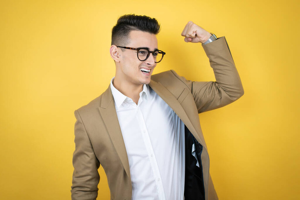 Mladý podnikatel přes izolované žluté pozadí ukazující paže svaly usmívající se hrdě - Fotografie, Obrázek