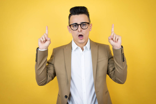 Young business man over isolated yellow background amazed and surprised looking at the camera and pointing up with fingers and raised arms - Photo, Image
