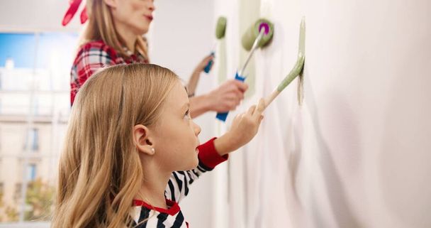 Side view. Close up of Caucasian cute little kid girl helping parents to paint wall in green color using roller brush. Mom talking to small daughter renovating and redesigning kid room. Family concept - Photo, Image