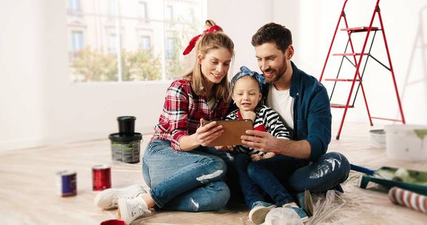 Happy Caucasian little adorable girl sitting with parents, tapping and browsing online on tablet, choosing new room decor together. home repair. Renovating and improving house. renovation concept - Foto, immagini