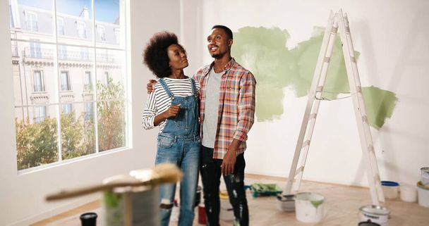 Happy young African American couple stands in room in new own apartment discussing design and decor of house. Wife talking to husband about home repair. Renovation and improving apartment concept - Фото, изображение
