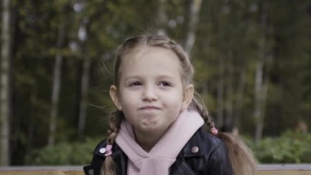 Porträt eines lustigen kleinen Mädchens mit Zöpfen, das verärgert oder frustriert auf verschwommenem grünen Baumhintergrund aussieht. Handeln. Konzept der Kindheit und Emotionen. - Filmmaterial, Video