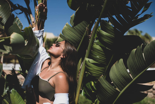 милая молодая девушка-подросток или женщина позирует с банановыми листьями дерева, одетые в летние хипстерские винтажные белые стильные одежды. Модная девушка позирует. Смешная и позитивная женщина в солнечных очках - Фото, изображение