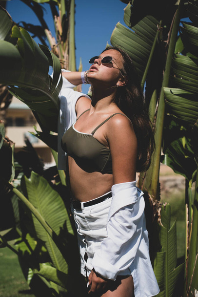 cute young teenage girl or woman posing with banana tree leaves dressed in summer hipster vintage white stylish clothes. Trendy girl posing. Funny and positive woman in sunglasses - Fotó, kép