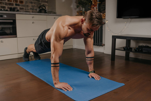 Ein muskulöser Mann macht Liegestütze auf einer blauen Yogamatte in seiner Wohnung. Ein athletischer Typ mit Tätowierungen auf den Unterarmen macht zu Hause ein Brusttraining. Ein Bodybuilder mit nacktem Oberkörper - Foto, Bild