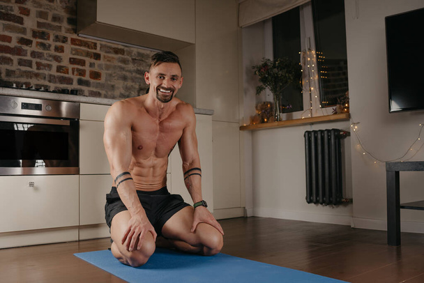 A muscular man is sitting on a blue yoga mat in his apartment. An athletic guy with tattoos on his forearms is relaxing between sets of pushups at home. A bodybuilder with a naked torso in the evening - Foto, afbeelding