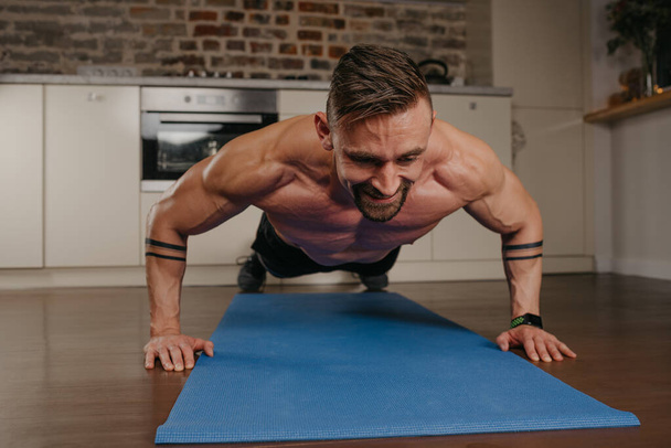 A happy muscular man with a beard is doing pushups on a blue yoga mat in his apartment in the evening. An athletic guy with tattoos on his forearms is doing a chest and triceps workout at home. - Photo, Image