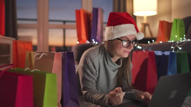 Onnellinen nainen silmälasien yllään joulupukin hattu makaa matolla ja tekee verkko-ostoksen luottokortilla ja kannettavalla tietokoneella. Ostoskassit. - Materiaali, video