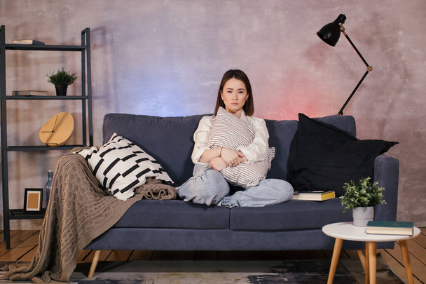Μια γυναίκα κάθεται σε έναν καναπέ σε ένα άνετο δωμάτιο βλέποντας τηλεόραση και αναστατωμένος από ό, τι βλέπει στην οθόνη - Φωτογραφία, εικόνα