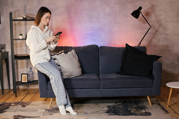 Jovem mulher asiática sentada em um sofá em um quarto acolhedor olha para o telefone - Foto, Imagem