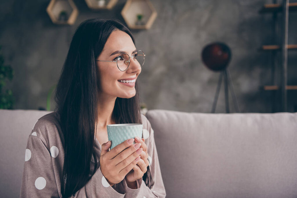 Profile portrait of nice optimistic girl hold cup look empty space wear spectacles pijama at home on sofa - Фото, изображение