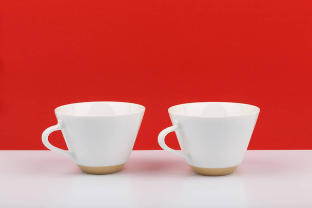 Two white ceramic coffee cups on white table against red background with space for text - Фото, изображение