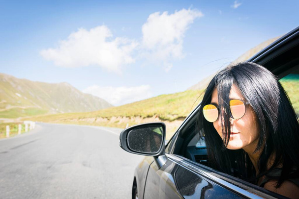 Mulher caucasiana de cabelos escuros picos rosto da janela do carro olhando direto para a câmera com asfalto estrada fundo - Foto, Imagem
