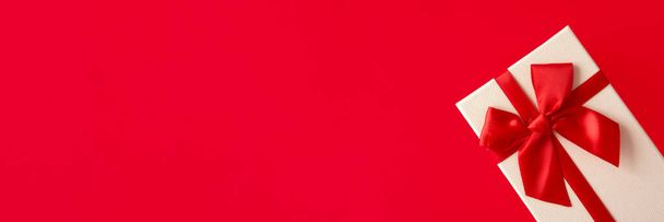 Bílý dárek s červenou stuhou na jasně červeném pozadí. Slavnostní pozadí pro dovolenou: Narozeniny, Valentýn, Vánoce, Nový rok. - Fotografie, Obrázek