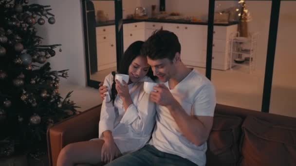 glückliches junges Paar trinkt Tee auf Sofa neben Weihnachtsbaum - Filmmaterial, Video