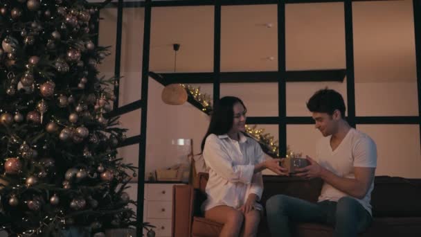 happy woman presenting gift to amazed boyfriend near christmas tree - Footage, Video