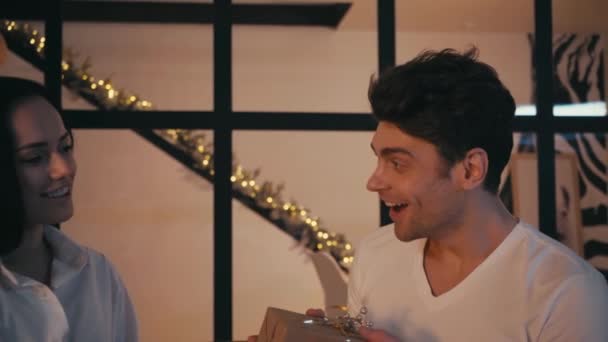 Frau überreicht erstaunten Freund Weihnachtsgeschenk und umarmt ihn auf Sofa - Filmmaterial, Video