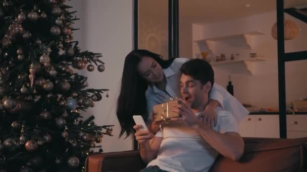 žena představuje dárek pro přítele chatování na smartphone v blízkosti vánočního stromečku - Záběry, video
