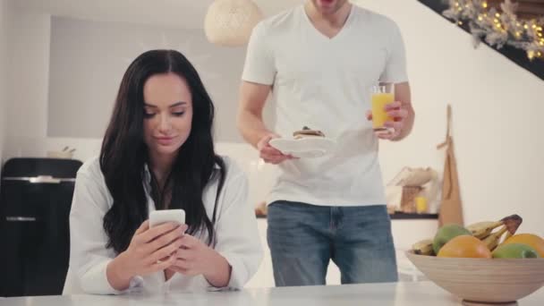 Nuori mies tarjoilee aamiaista lähellä onnellinen tyttöystävä chattailuun älypuhelimella - Materiaali, video
