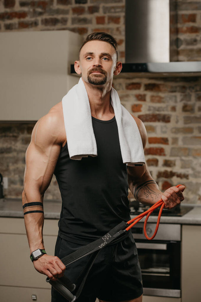 A muscular man with a white towel on the shoulders is posing with pull elastic rope in his apartment. The bodybuilder with tattoos on his forearms is demonstrating his sporty physique at home - Photo, Image