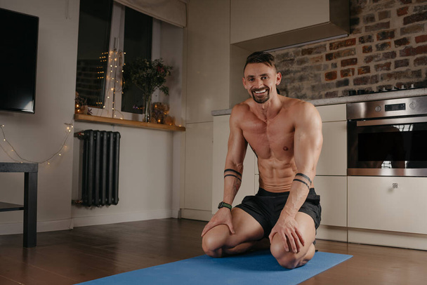 Muskularny mężczyzna siedzi na niebieskiej macie do jogi w swoim mieszkaniu. Sportowiec z tatuażami na przedramionach relaksuje się między zestawami pompek w domu. Kulturysta z nagim tułowiem wieczorem - Zdjęcie, obraz