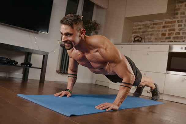 Ein muskulöser Mann macht Liegestütze auf einer blauen Yogamatte in seiner Wohnung. Ein athletischer Typ mit Tätowierungen auf den Unterarmen macht zu Hause ein Brust- und Trizepstraining. Ein glücklicher Bodybuilder mit nacktem Oberkörper - Foto, Bild