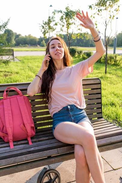 woman in city in summer sits bench, saw familiar friends, waving to her friends guys, holding smartphone, making phone call, rest after work, pink backpack, casual clothes. - Valokuva, kuva