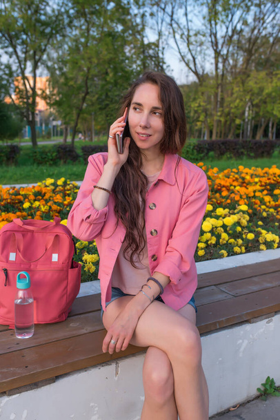woman city in summer sits bench, listens to voice message online chat application and Internet, holds a smartphone, rest after work, calls by phone, pink jacket, shorts, casual clothes. - Фото, изображение