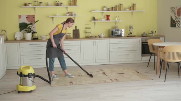Młoda kobieta z odkurzaczem odkurzania dywan w kuchni - Materiał filmowy, wideo