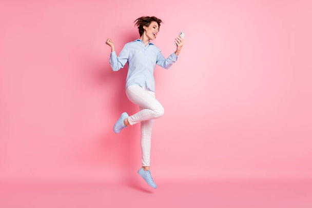 Full size profile photo of pretty lady good mood jump high up air student browsing telephone chatting friends celebrate weekend wear blue shirt pants shoes isolated pink color background - Zdjęcie, obraz
