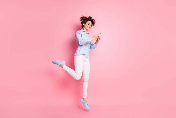 Full size profile photo of pretty lady good mood jump high up air carefree student browsing telephone chatting friends wear blue shirt white pants shoes isolated pink color background - Zdjęcie, obraz