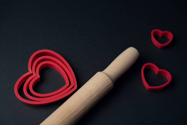 Red heart-shaped baking tins for cookies and wooden rolling pin on the dark (black) background. Flat lay, top view, space for text. Valentine's day concept. - Foto, Imagen