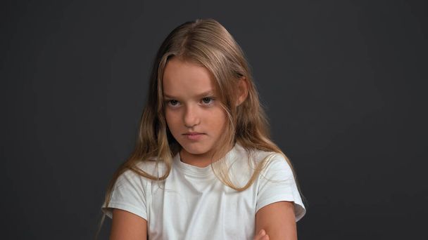 Disappointment on the face of a lovely baby little girl with hands folded, looking at the camera wearing white t-shirt and black pants isolated on black background - Фото, изображение
