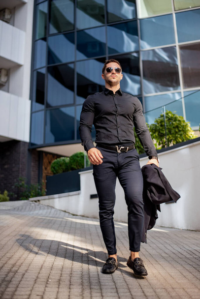 Retrato de un hermoso hombre hipster musculoso con camisa negra y gafas de sol posando en las calles de la ciudad europea - Foto, imagen