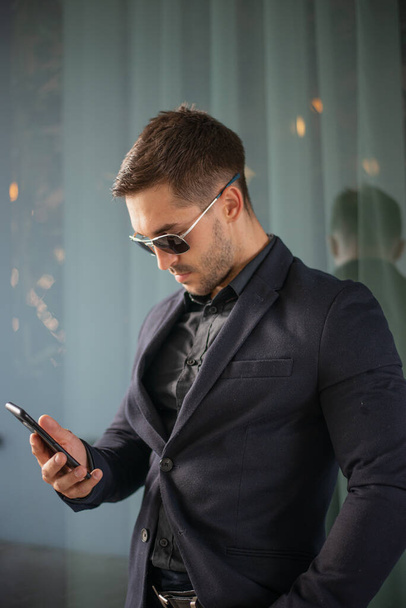 Κομψός όμορφος άντρας με γενειάδα, φοράει σακάκι κοστούμι και πουκάμισο, έξω στο δρόμο της πόλης. Άνθρωπος που χρησιμοποιεί κινητό τηλέφωνο. Λεπτομέρειες του κλασικού κομψό επίσημο ένδυμα των ανδρών. - Φωτογραφία, εικόνα