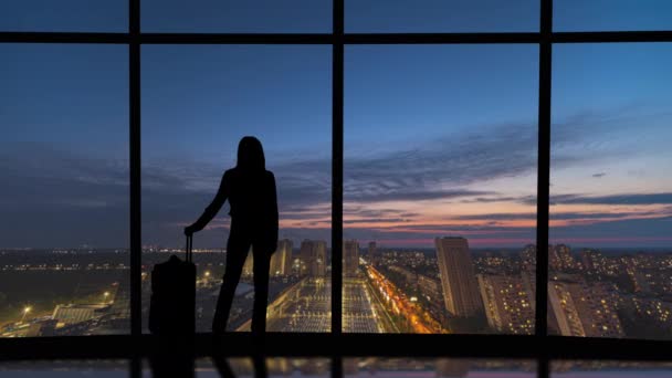 Die Frau mit dem Koffer steht neben den Fenstern mit nächtlichem Blick auf die Stadt. Zeitraffer - Filmmaterial, Video