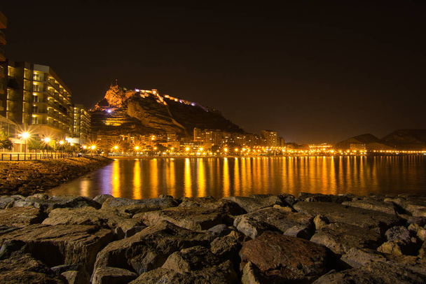 plaża u podnóża zamku z odbiciami światła miasta w morzu, w Hiszpanii - Zdjęcie, obraz