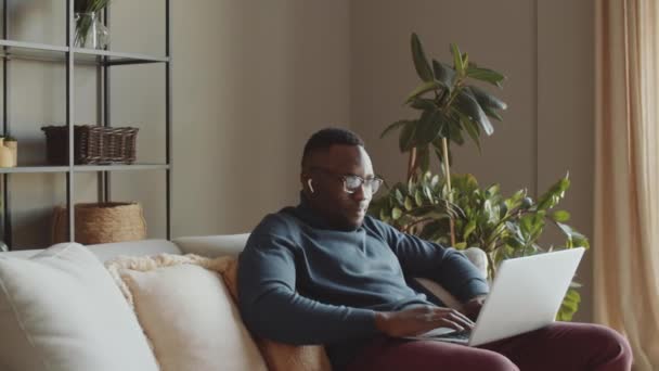 Kablosuz kulaklıklı genç Afro-Amerikalı adam oturma odasında kanepede oturuyor ve dizüstü bilgisayarından online video görüşmesi yapıyor. - Video, Çekim