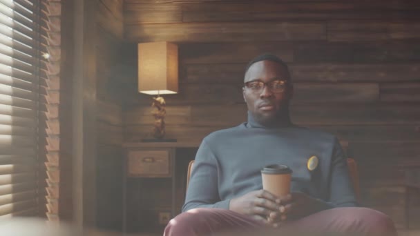 Portret van de jonge Afro-Amerikaanse man in slimme casual outfit en bril zittend op stoel, wegwerpkoffiekopje vasthoudend en poserend voor camera met vertrouwen - Video